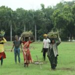 A group of Indian farmers working in lush green fields, carrying baskets of grass on their heads.