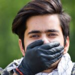 Close-up of a man outdoors with a black latex glove covering his mouth, conveying silence.