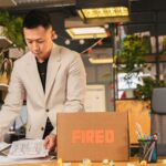 An employee packing documents into a box labeled 'FIRED' in an office environment.