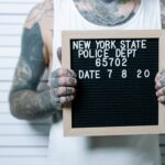 Close-up of tattooed man holding a mugshot sign in front of a height chart.