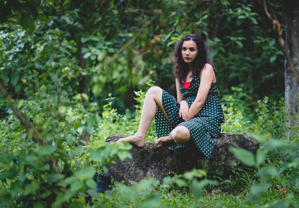 Woman in a green polka dot dress sitting on a rock in a lush forest setting.