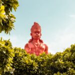 A vibrant red statue of Hanuman surrounded by green foliage under a clear sky.