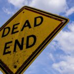 Yellow dead end sign against a blue sky with clouds, symbolizing stop or warning.