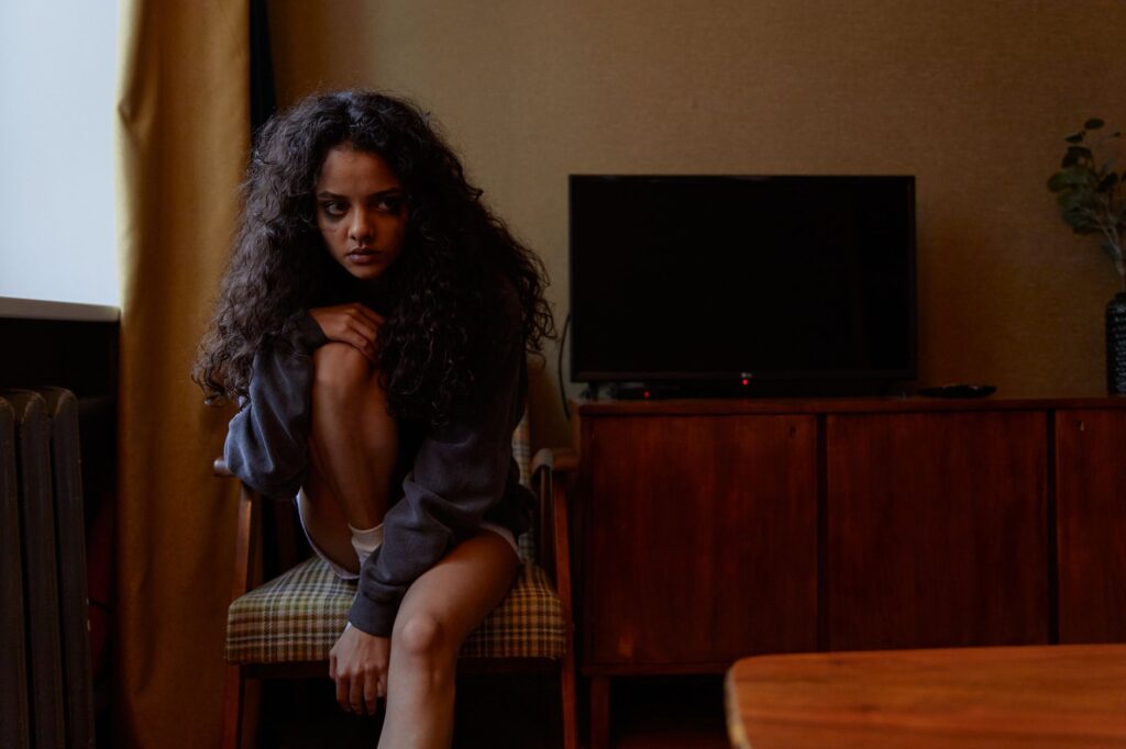 A woman sits contemplatively indoors, expressing deep emotion in a dimly lit room.