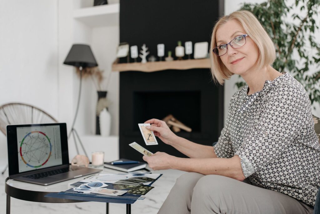 Woman with tarot cards using laptop for astrology reading in cozy living room.