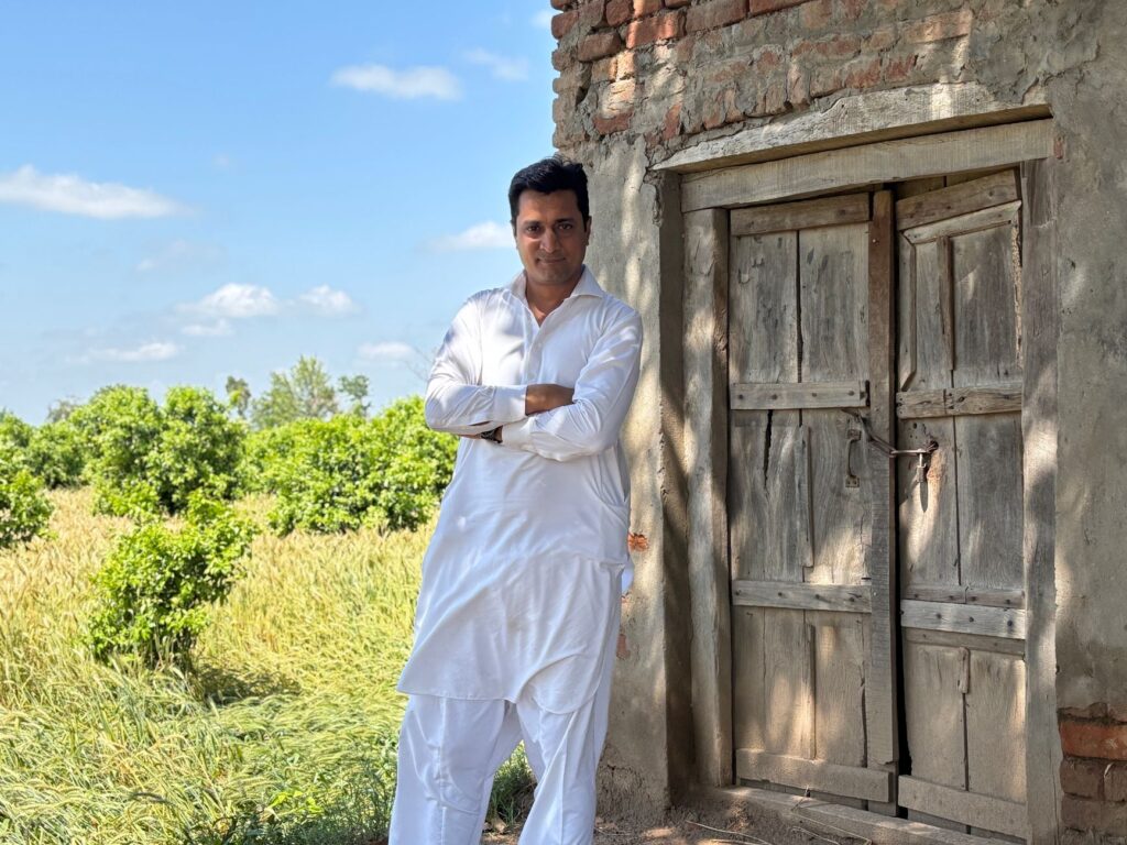 Man in traditional white attire stands confidently by an old wooden door in a rural setting, under a clear blue sky.
