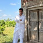 Man in traditional white attire stands confidently by an old wooden door in a rural setting, under a clear blue sky.