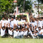 A joyful group of young men celebrating outdoors in a park during summer with a sign and white t-shirts.
