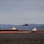 A helicopter flies over several cargo ships in a vast ocean with a mountainous backdrop.