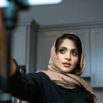 Woman in hijab recording a video on smartphone indoors, focusing on social media content creation.