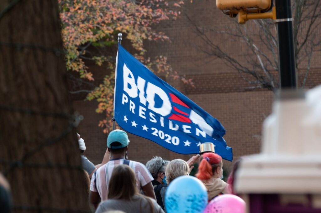 A diverse crowd gathered outdoors holding a Biden 2020 flag during a protest.