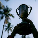 Silhouetted hands raise a trophy against a tropical palm tree backdrop, symbolizing victory.