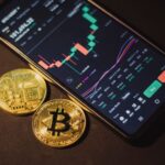 Smartphone displaying trading app with Bitcoin coins on a dark surface.