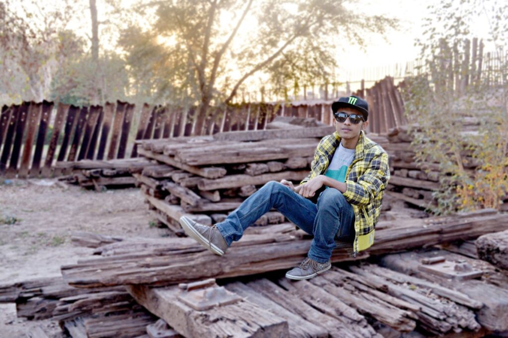 Casual young man in checkered shirt and cap sitting on stacked logs outdoors.