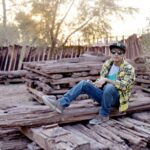 Casual young man in checkered shirt and cap sitting on stacked logs outdoors.
