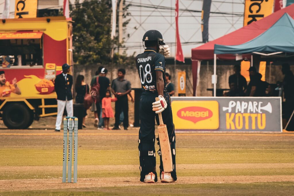 A cricket player stands ready on the field during a sunny match, surrounded by spectators.