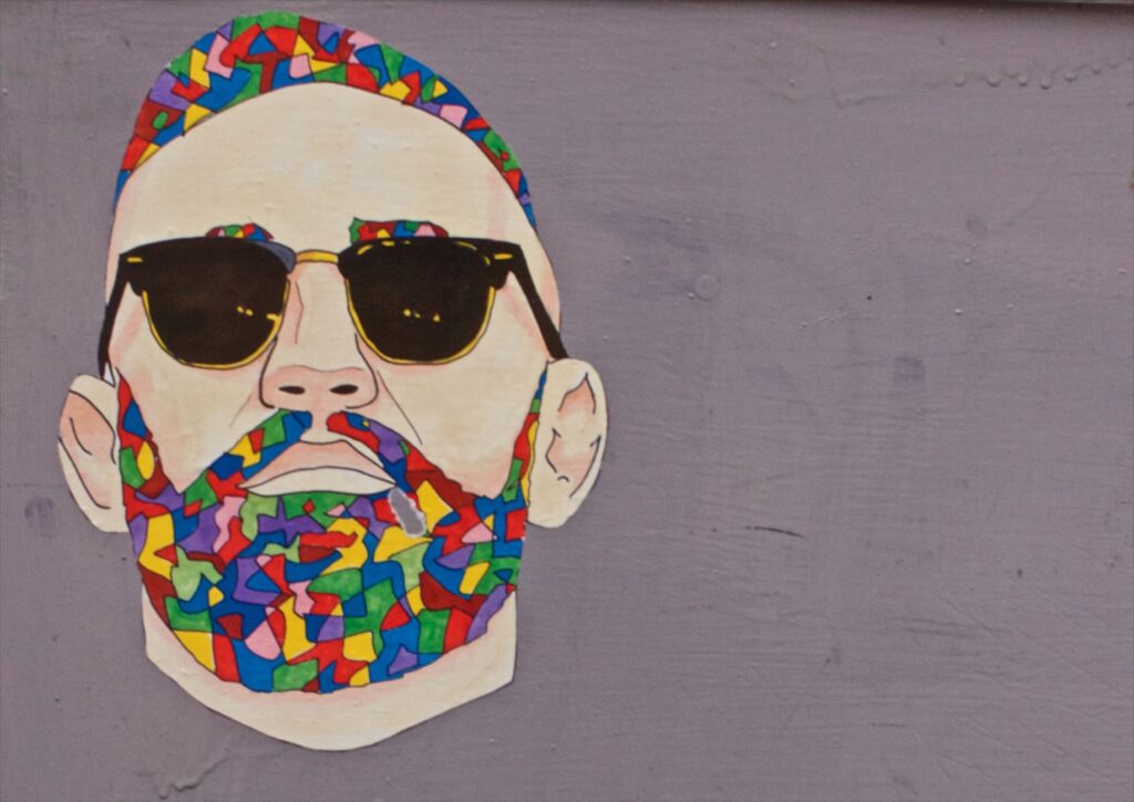 Abstract street art portrait of a man with colorful beard and sunglasses on a grey wall.
