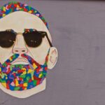 Abstract street art portrait of a man with colorful beard and sunglasses on a grey wall.