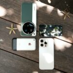 Assorted smartphones displayed outdoors on a wooden table under dappled sunlight.