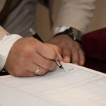 A close-up of a man signing a document, showcasing a wedding ring and pen.