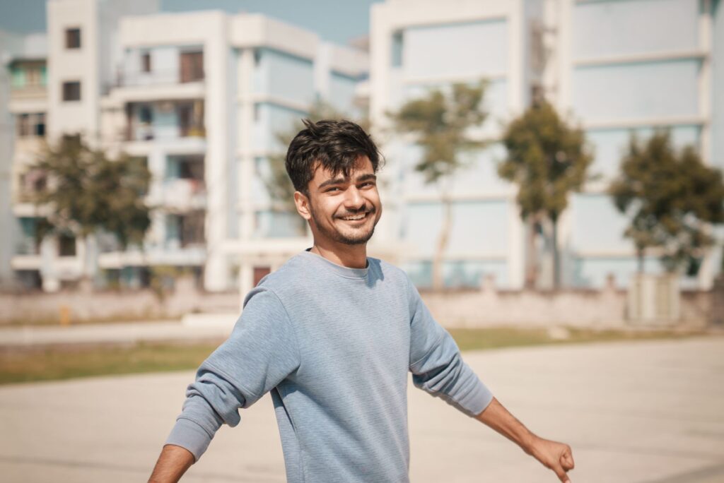Young man outdoors with a smile, wearing a blue sweater with buildings in the background.