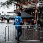 A man in a sports jersey walks through a rainy city street, navigating wet pavement and barriers.