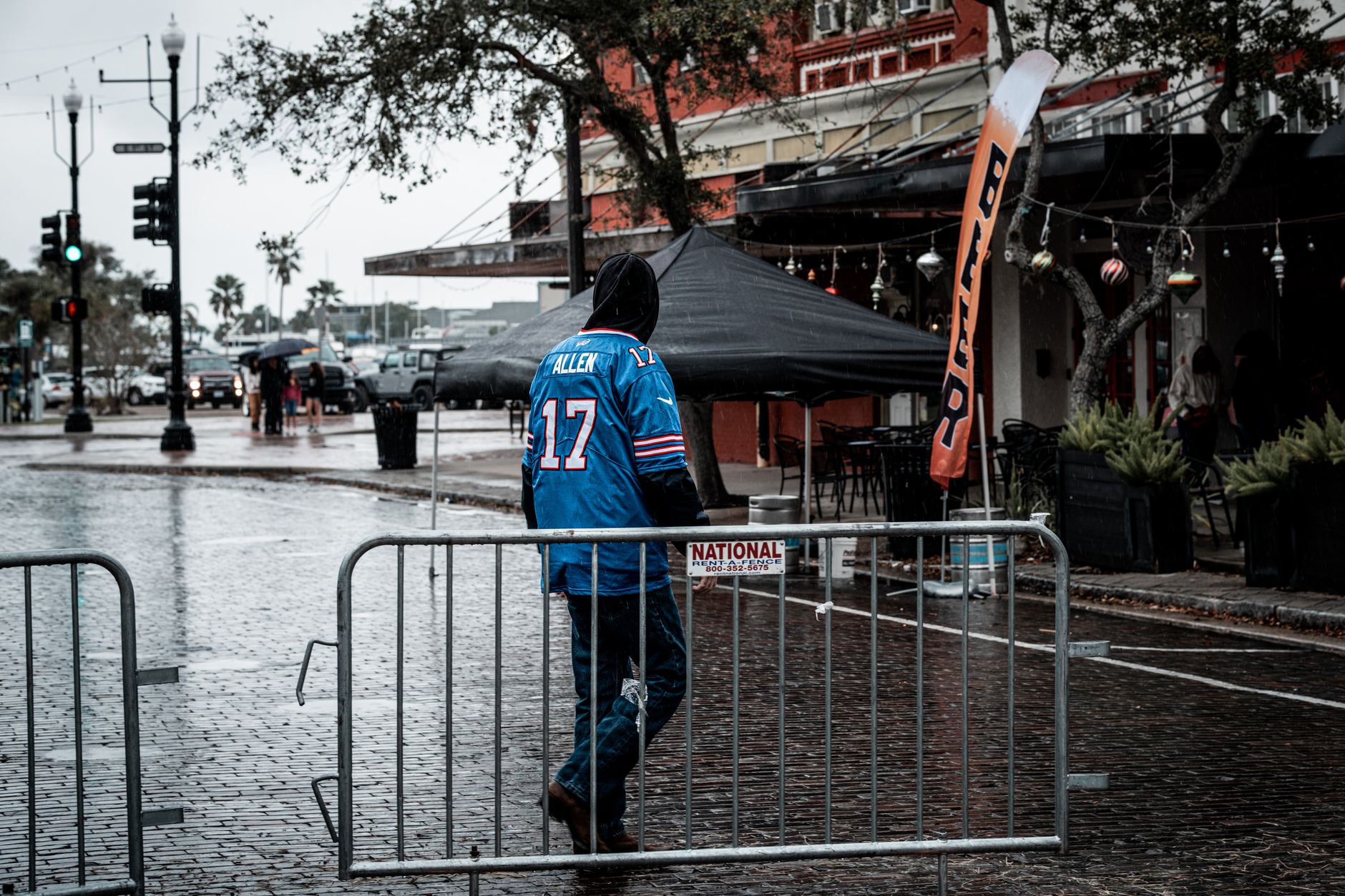 A man in a sports jersey walks through a rainy city street, navigating wet pavement and barriers.