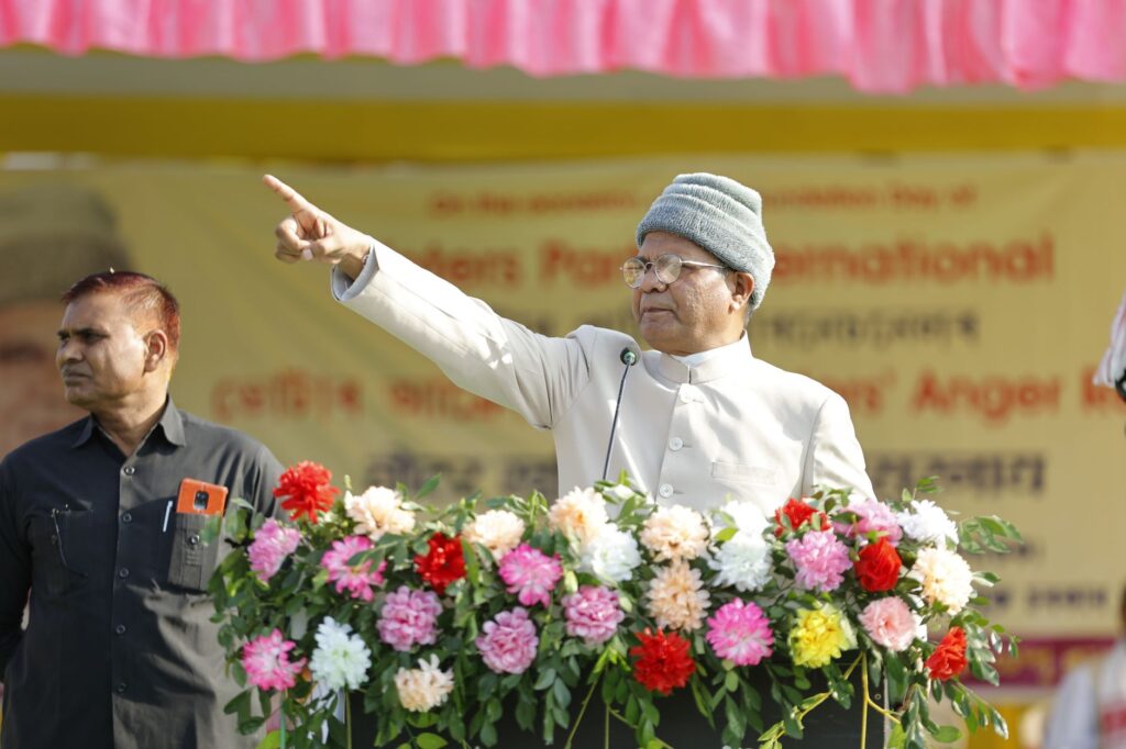Elderly man in traditional attire giving a speech outdoors, surrounded by flowers.