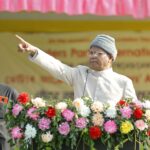 Elderly man in traditional attire giving a speech outdoors, surrounded by flowers.