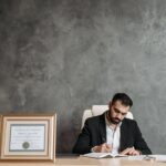 A business professional writing at a desk with a framed certificate and justice statue.