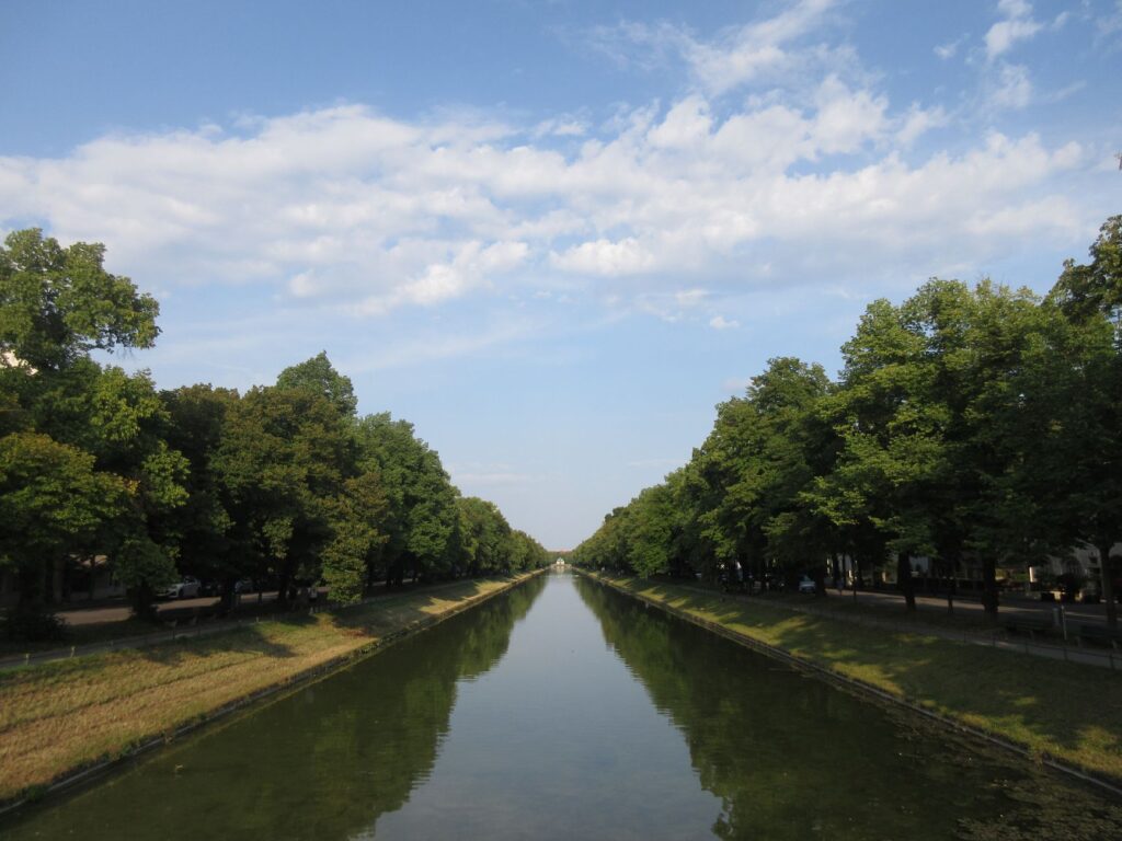 Tranquil water canal in Munich with reflections, lined by lush trees under a vibrant sky.