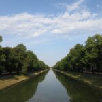 Tranquil water canal in Munich with reflections, lined by lush trees under a vibrant sky.