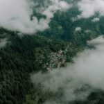 Aerial photograph showcasing a small village nestled among lush green mountains and clouds in Himachal Pradesh, India.