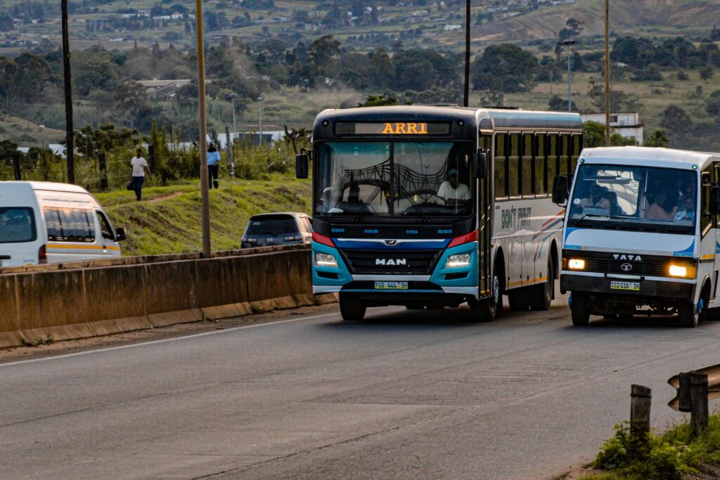 Street scene showing buses on a road with hilly landscape in the background.