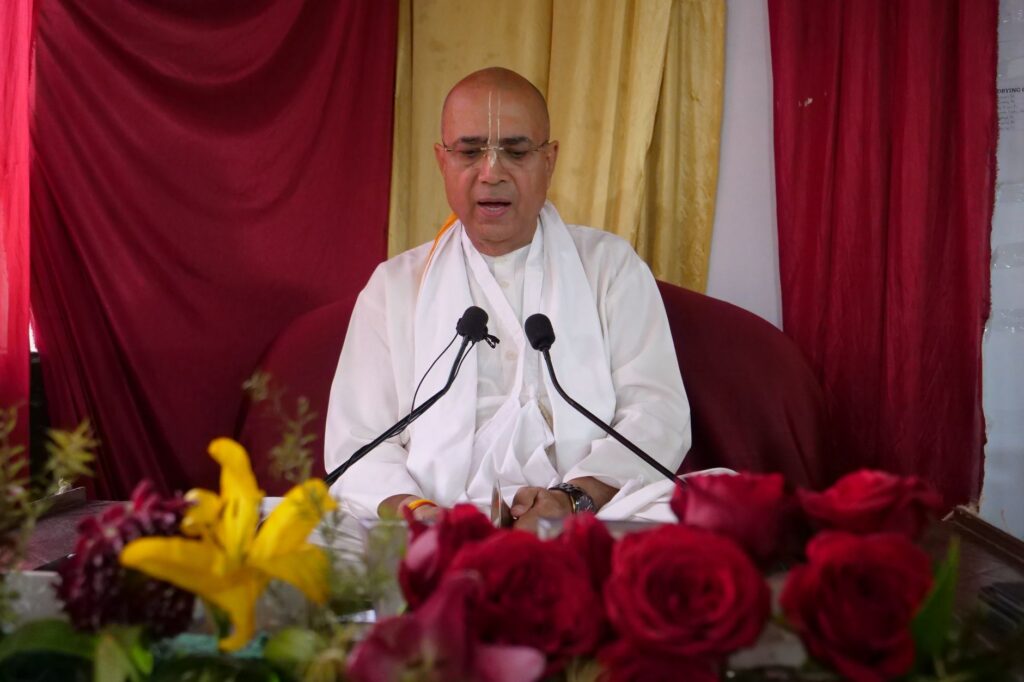A senior spiritual leader delivers a religious sermon surrounded by colorful flowers and microphones.