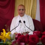 A senior spiritual leader delivers a religious sermon surrounded by colorful flowers and microphones.