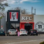 Street view of a KFC restaurant in Manzini, Eswatini, showing parked cars and patrons entering.