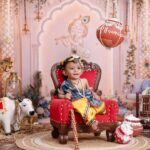 Adorable baby dressed as Lord Krishna surrounded by traditional festive decor and colorful pottery.