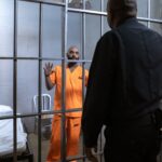 A male prisoner in an orange uniform stands behind bars facing a guard in a jail cell.