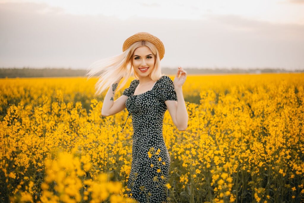 Woman in sundress and hat smiling amidst vibrant yellow flowers in a sunny field.
