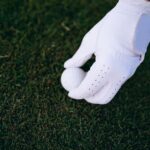 Close-up of a golfer's hand in a glove placing a golf ball on a green field.