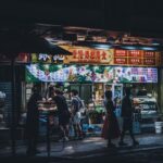 A bustling urban street food stall illuminated at night with passersby enjoying snacks.