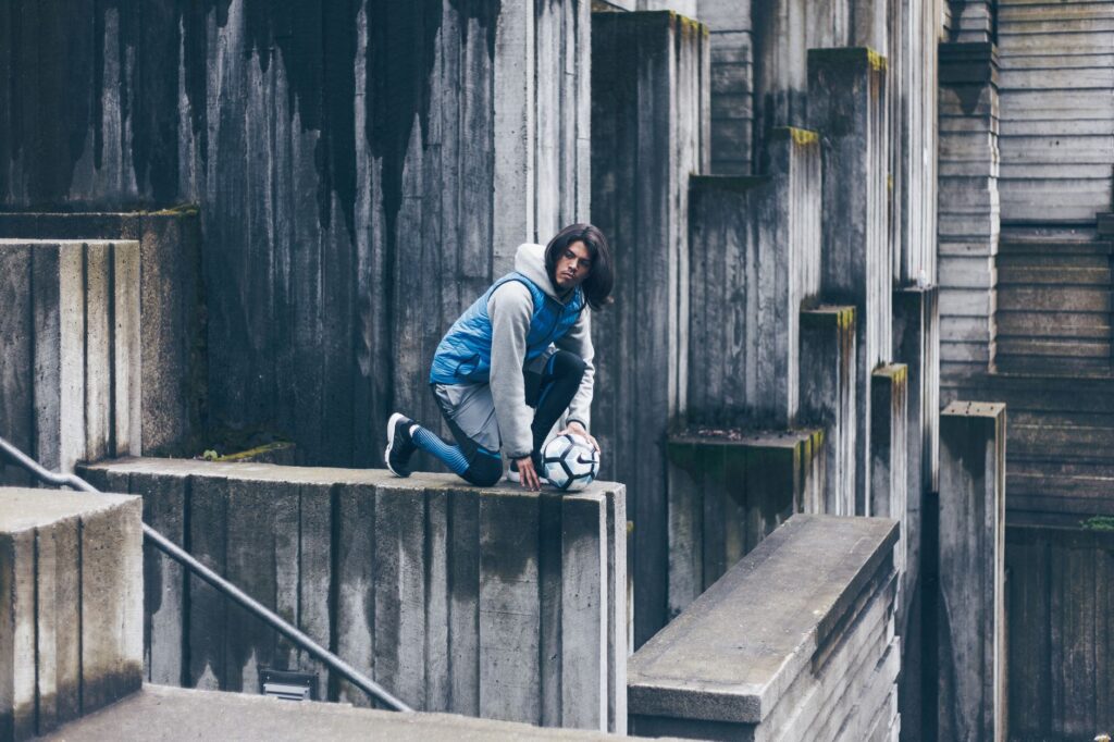 Side view of concentrated male footballer sitting with ball on stone wall in city and looking away