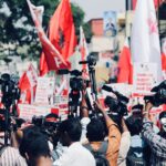 A vibrant protest in Chennai, India with media coverage featuring bright red flags and banners.