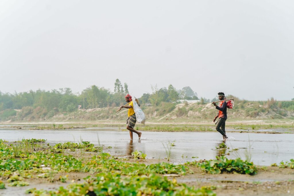 Two men crossing a shallow river with greenery around, under a clear sky.