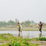 Two men crossing a shallow river with greenery around, under a clear sky.