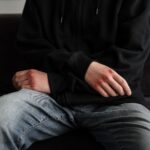 A close-up of a teenager with bruised hands, wearing a hoodie and jeans, sitting on a couch.