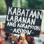 A protester holds a sign against corruption in a urban street setting.