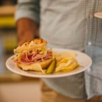 Plate with a tasty sandwich, chips, and a pickle held by a man indoors.