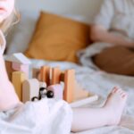 Crop faceless toddler girl in casual wear sitting on bed and playing with colorful wooden blocks while mother doing remote work with laptop on knees on background